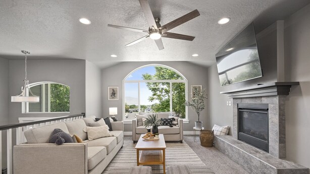 A modern living room with a fireplace, a ceiling fan, and a large window offering a view of trees.