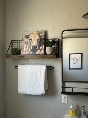 A bathroom with a towel hanging on a rack, a shelf with a plant and a painting, and a mirror with a framed picture. - Millz Manor at Fall River Millz (Fall River Mills)
