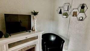 Un soggiorno con una TV a schermo piatto su un camino bianco, una poltrona in pelle nera, un camino acceso e decorazioni murali esagonali.