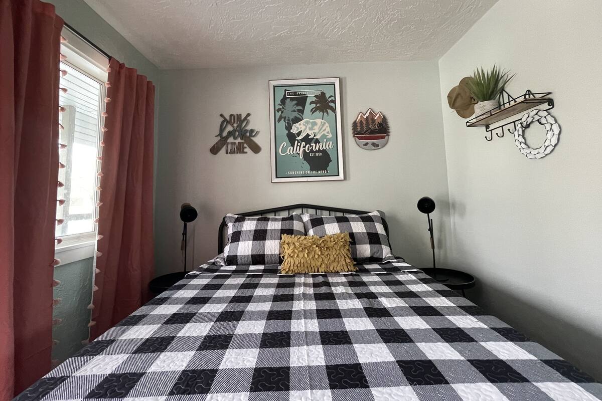 A bedroom with a checkered bedspread, a framed California poster, decorative wall art, and a plant on a shelf.