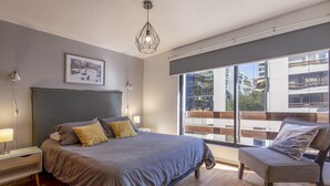 A modern bedroom with a large bed, a nightstand, a reading lamp, a chair, and a large window offering a view of buildings.