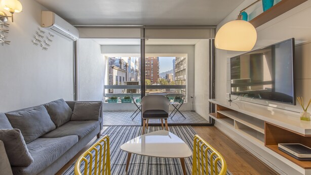 A modern living room with a grey sofa, a white coffee table, and a TV on a white stand. There is a balcony with a view of buildings.