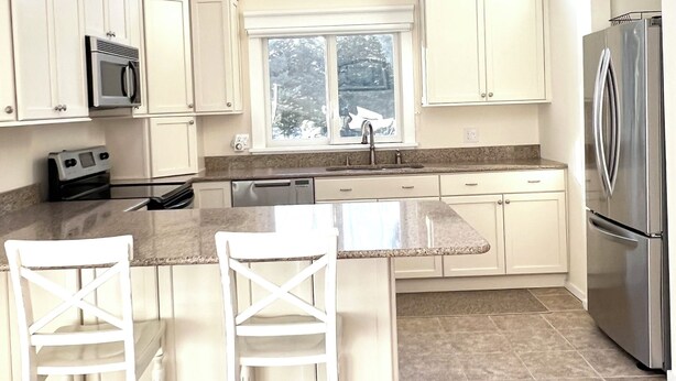 A modern kitchen with white cabinetry, a stainless steel refrigerator, a microwave, and a large window above the sink. - Pristine Remsenburg Retreat to Play & Work All Summer Long (Remsenburg)