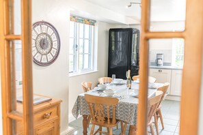 A dining area with a table set for four, chairs, a clock on the wall, and a kitchen in the background. - Middlefield Cottage, rural location close to lake (Coniston)