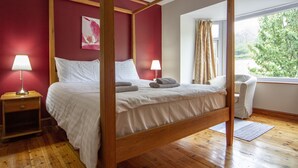 Ein Himmelbett mit weißen Leinen, ein Nachttisch aus Holz mit einer Lampe und Blick auf Grün durch ein Fenster.