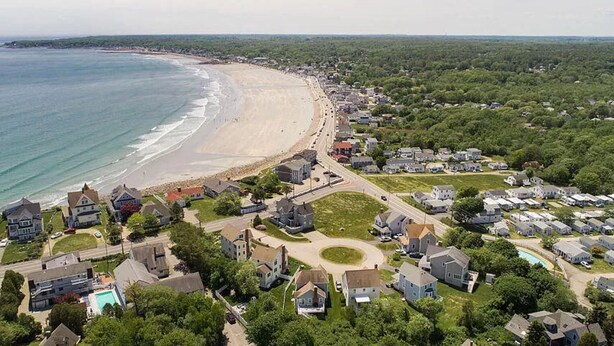 Vue aérienne d’un quartier résidentiel situé sur la côte, avec des maisons, une plage et une petite piscine.