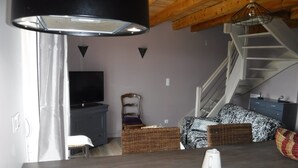 A modern living room with a flat-screen TV, a dining area with a table and chairs, a staircase, and a kitchen area with a stove.