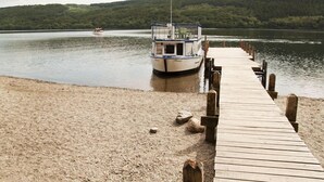 A wooden pier leading to a boat docked on a lake with a forested hill in the background. - Mill Pool Barn, stunning riverside barn conversation in idyllic location (Coniston)