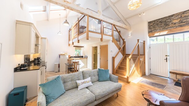 A spacious living area with a high ceiling, a staircase leading to an upper floor, and a kitchen area. - Mill Pool Barn, stunning riverside barn conversation in idyllic location (Coniston)