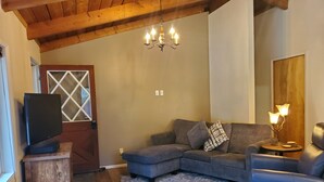 Un soggiorno con un divano componibile, una poltrona blu, un tavolino in legno, una TV su un mobile in legno, un lampadario e una porta con un motivo geometrico in vetro.