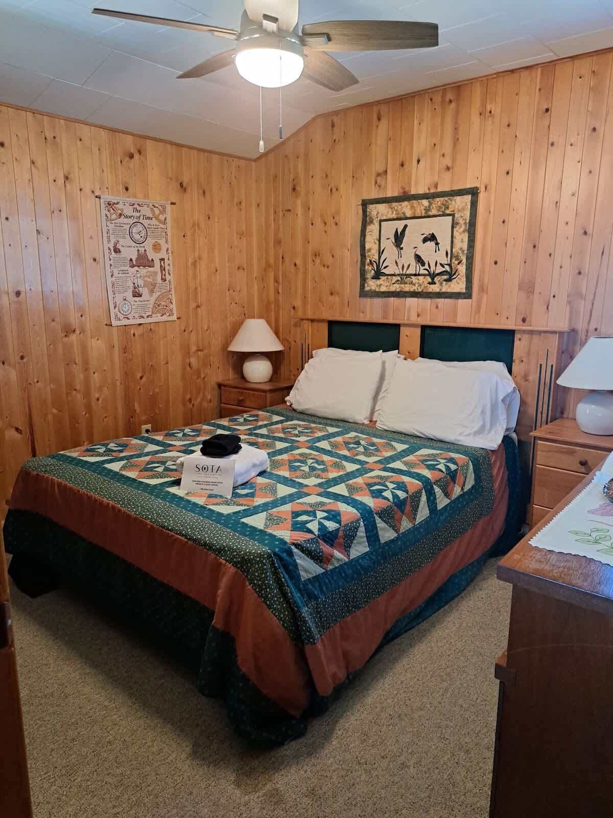 Una habitación con paneles de madera, una cama, un ventilador de techo, una mesita de noche, una lámpara y un cuadro en la pared.