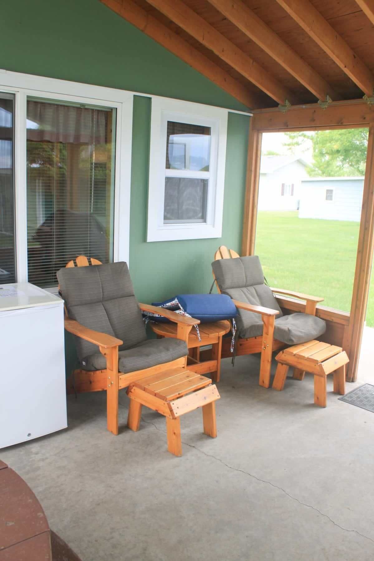 A covered patio area with two wooden chairs and a small table, a white refrigerator, and a window with blinds.