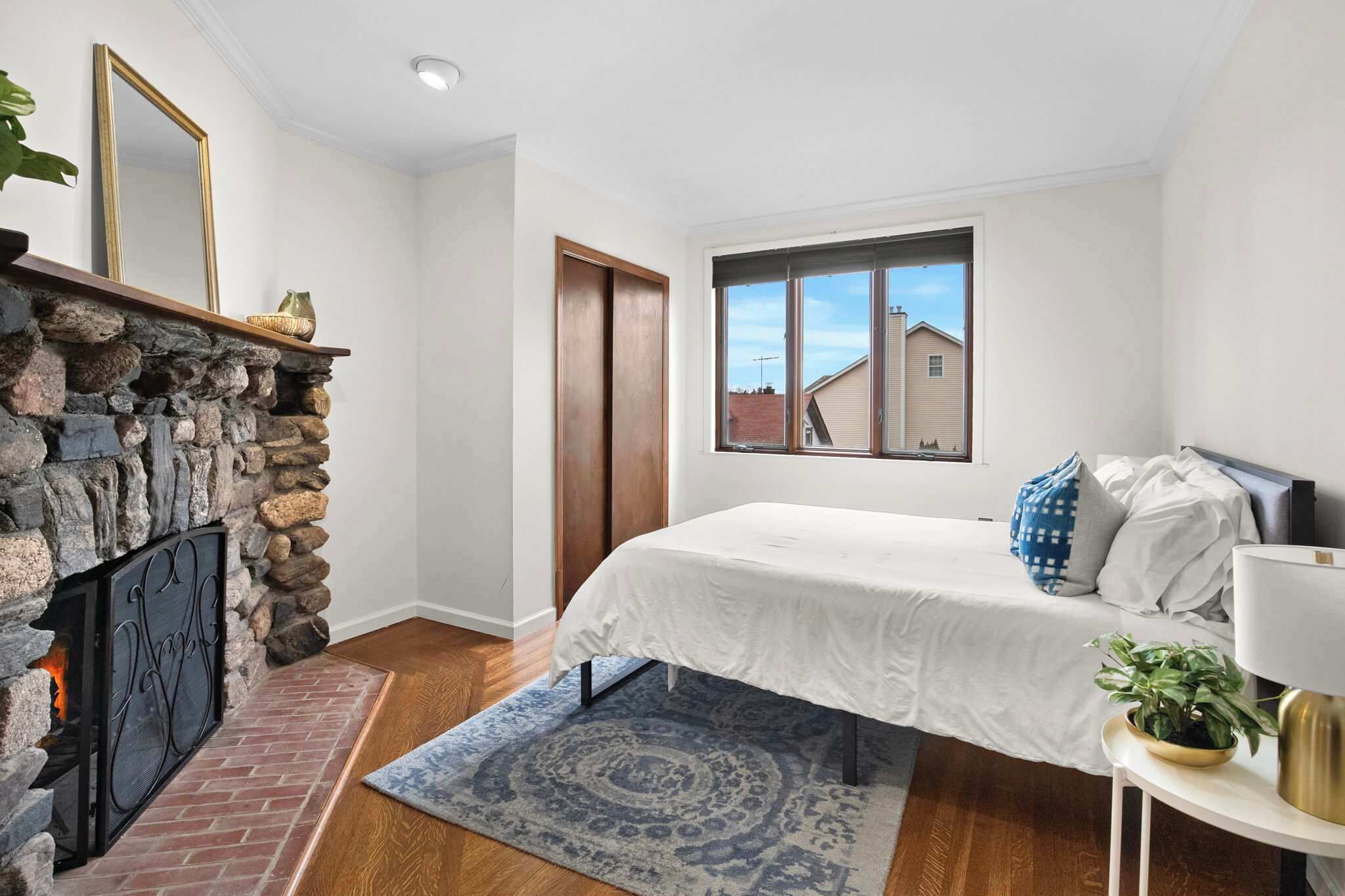 A bedroom with a stone fireplace, a bed with white bedding, a window with a view of houses, and a small table with a lamp.