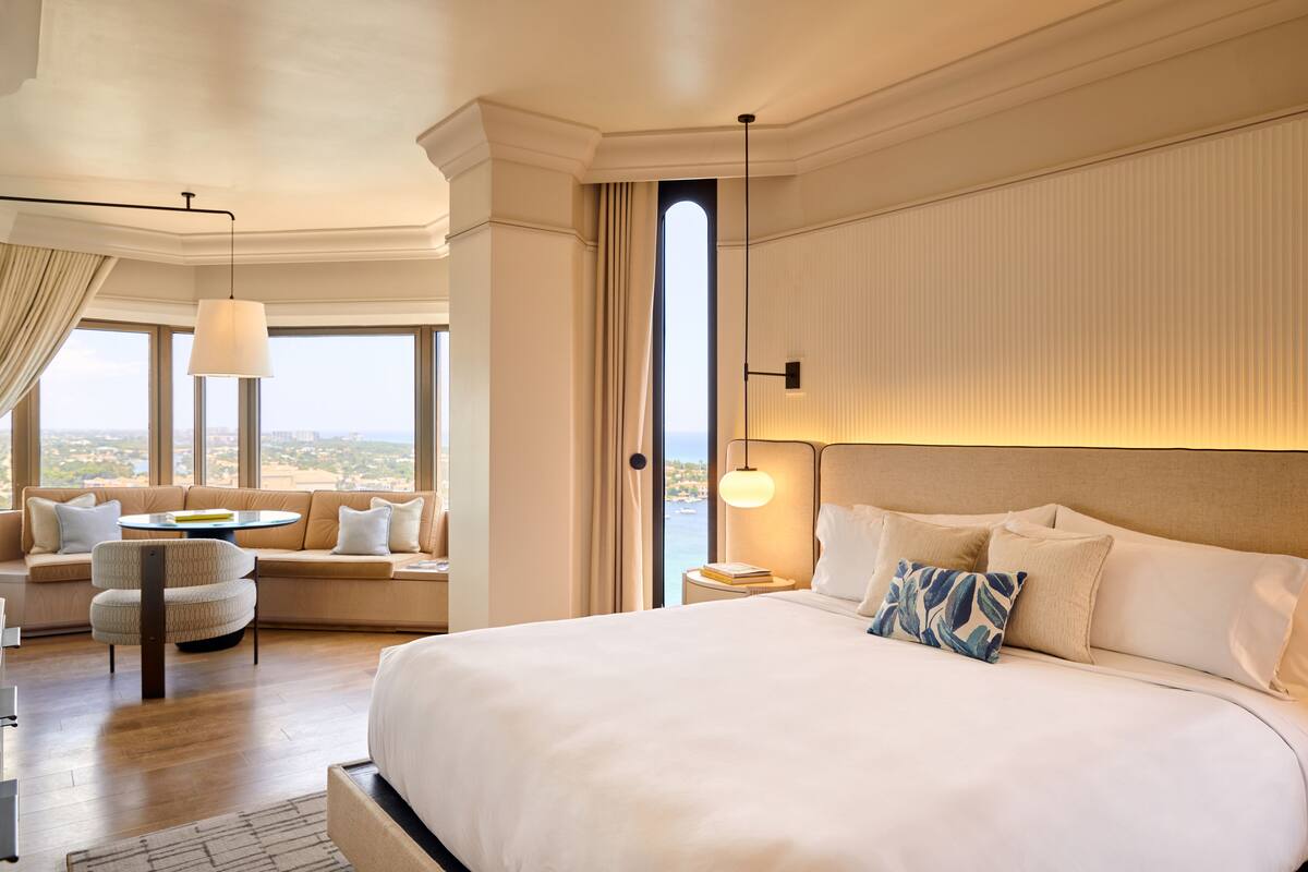 A spacious bedroom with a large bed, a round coffee table, a sofa, and a view of the city.