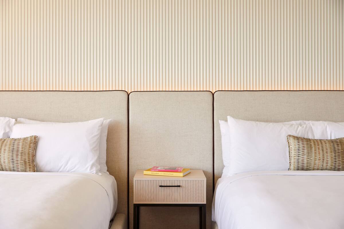 A hotel room with two beds, each with white linens and a woven pillow. A small bedside table with a book and a drawer is centered between the beds.