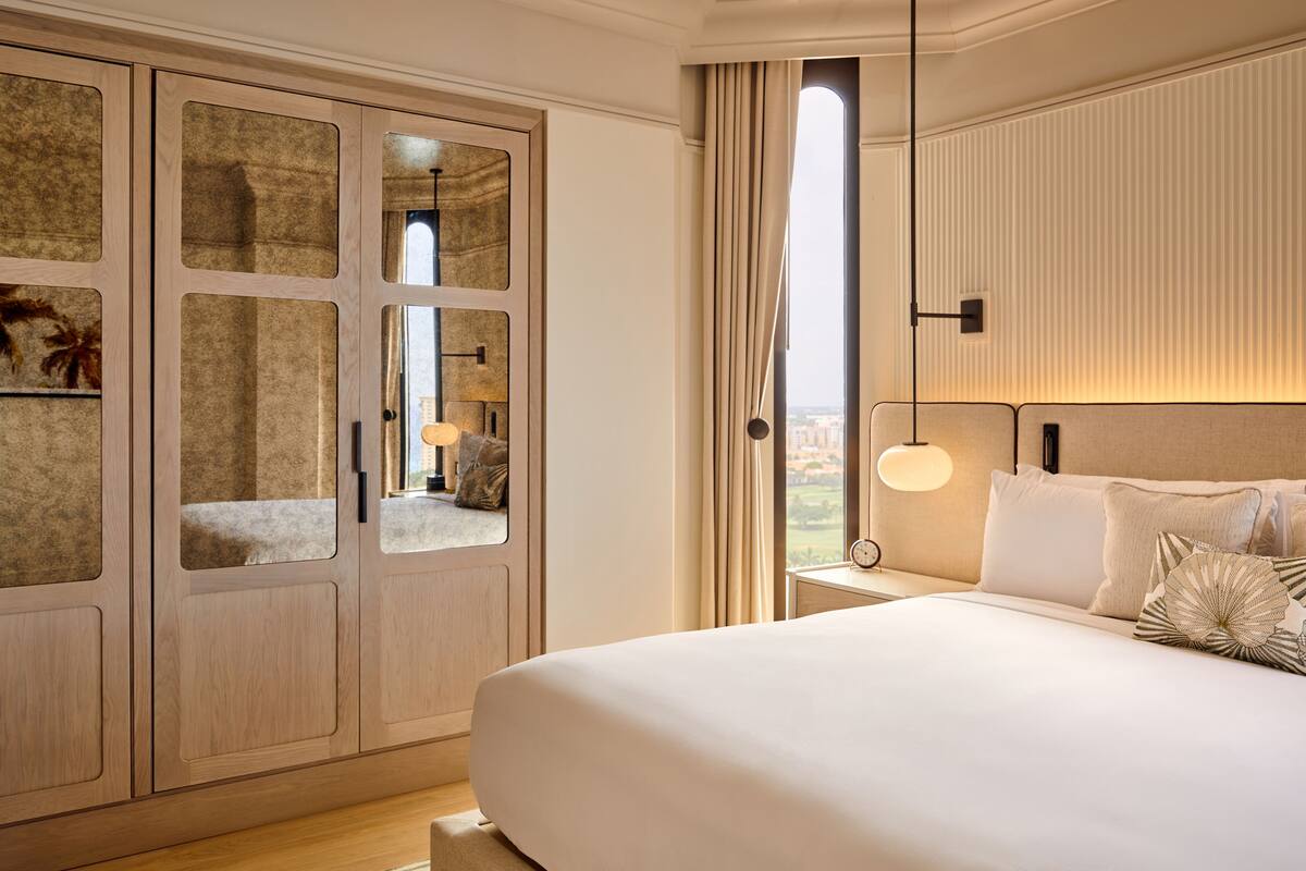 A hotel room with a large bed, a wardrobe with mirrored doors, a nightstand with a lamp, and a view of the city through the window.