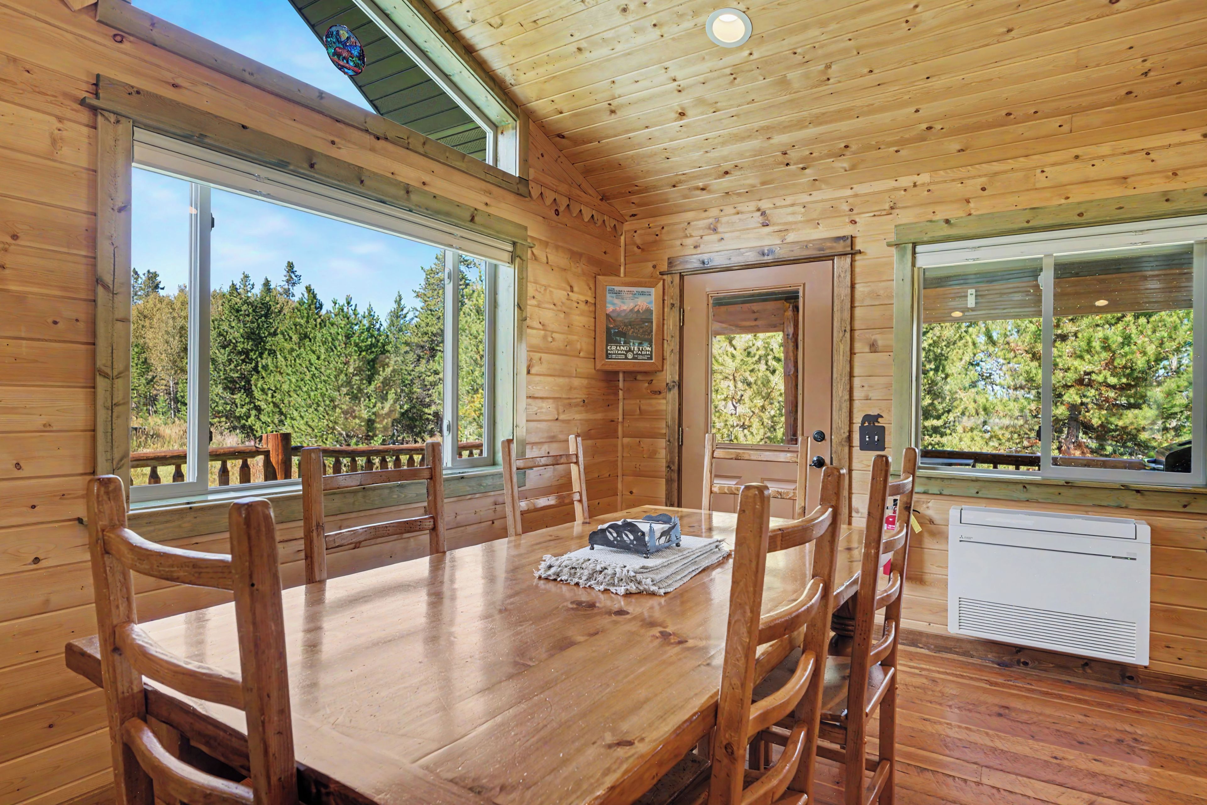 A wooden cabin interior with a dining table, chairs, and large windows offering views of a forest.