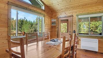 A wooden cabin interior with a dining table, chairs, and large windows offering views of a forest.