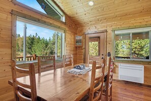 Interior de una cabaña de madera con una mesa de comedor, sillas y ventanas amplias que dan vistas a un bosque.