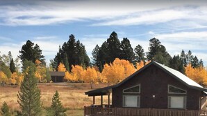 A cabin with a deck, surrounded by autumn foliage and a grassy field.