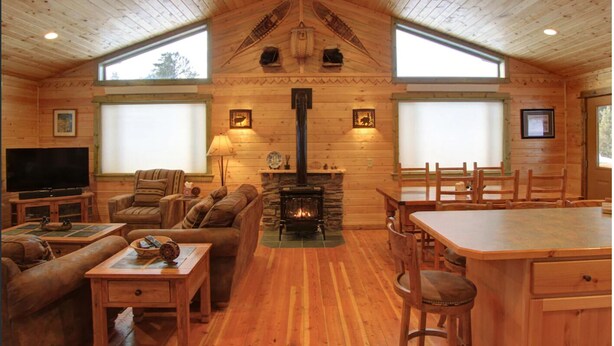 A log cabin interior with a fireplace, wooden furniture, and large windows.