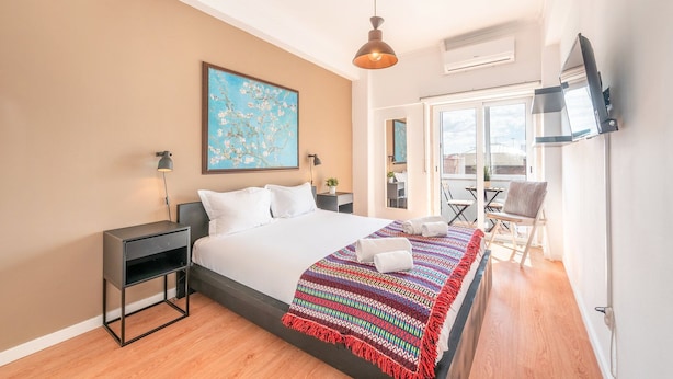 Une chambre moderne avec un grand lit, une couverture colorée, un tableau, un bureau et un balcon avec des chaises.