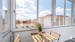 Un balcon avec une table et des chaises en bois, une plante en pot et une vue sur les immeubles à travers de grandes fenêtres.