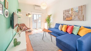 Un salon avec un canapé bleu, des coussins colorés, une table basse en bois, un tapis à motifs, un fauteuil suspendu et un grand tableau accroché au mur.