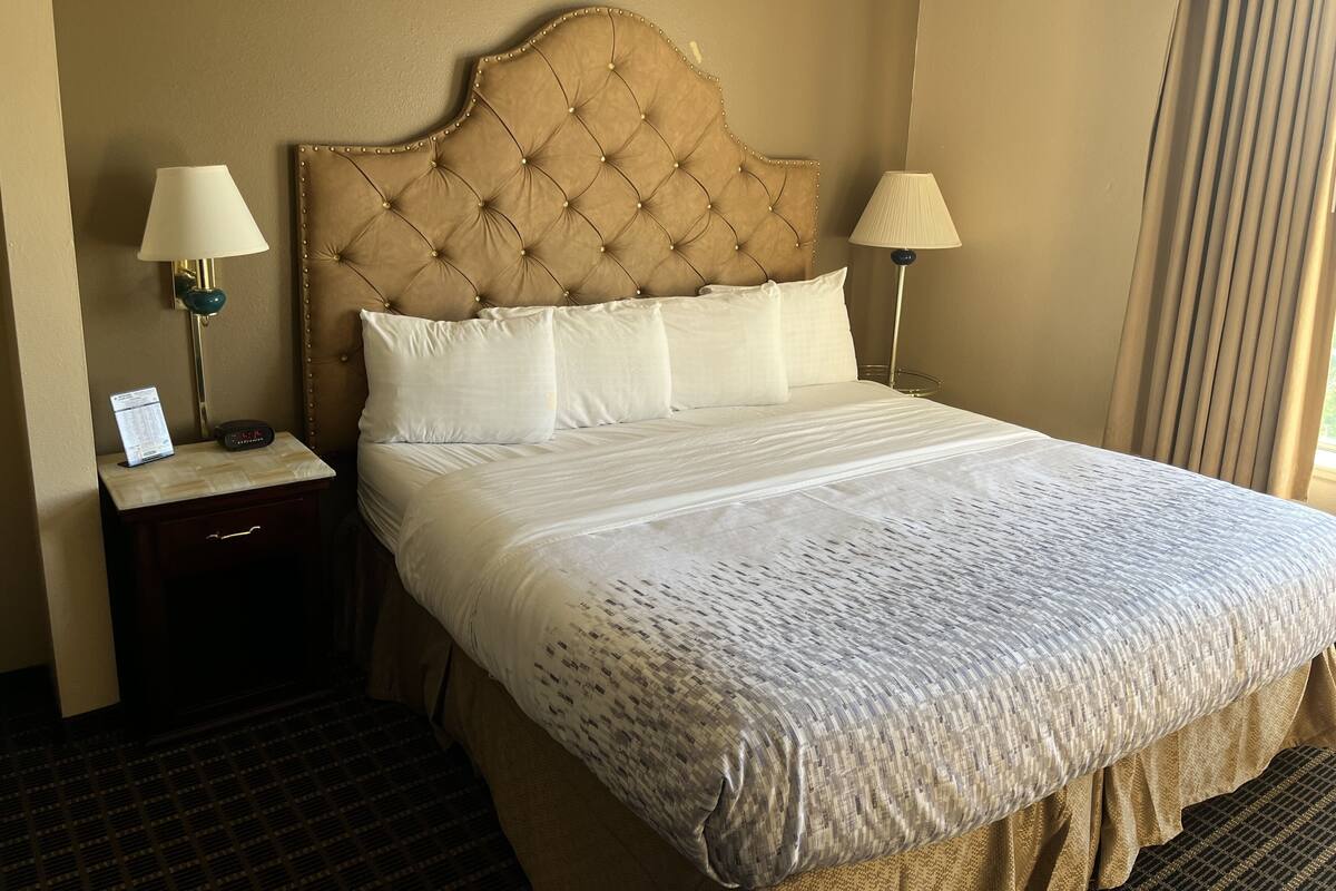 A hotel room with a tufted headboard, white bedding, two bedside lamps, a nightstand with a lamp and a clock, and a window with curtains.
