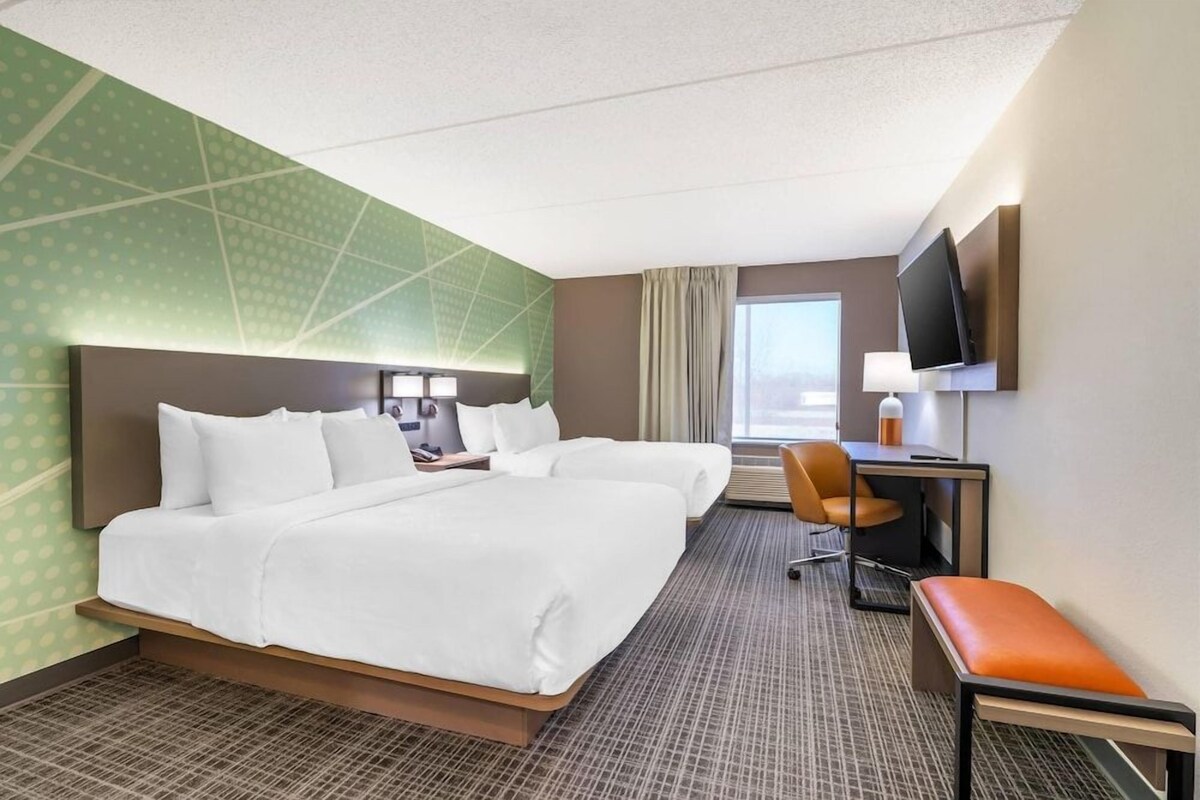 A hotel room with two beds, a desk, a chair, a TV, and a window.