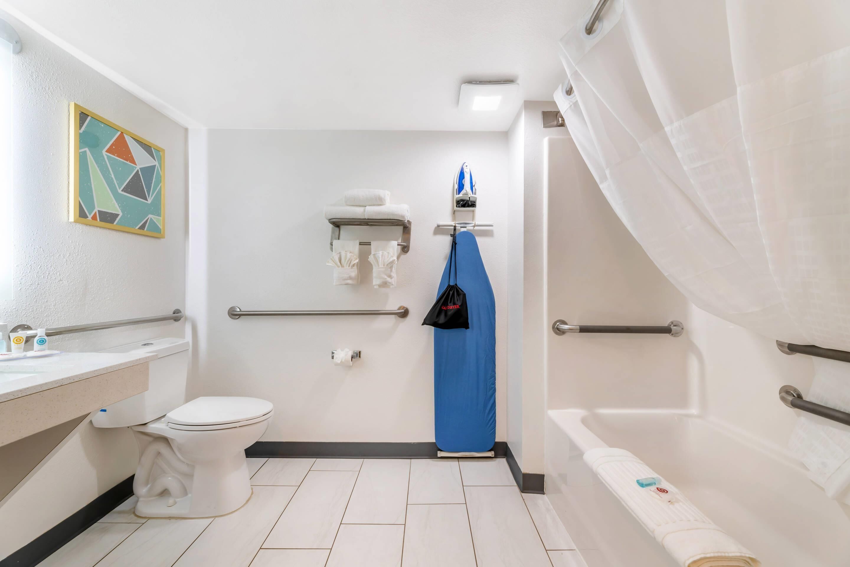 Un baño con inodoro, lavamanos, ducha y bañera. Hay barras de apoyo, un perchero para toallas y una cortina de ducha.