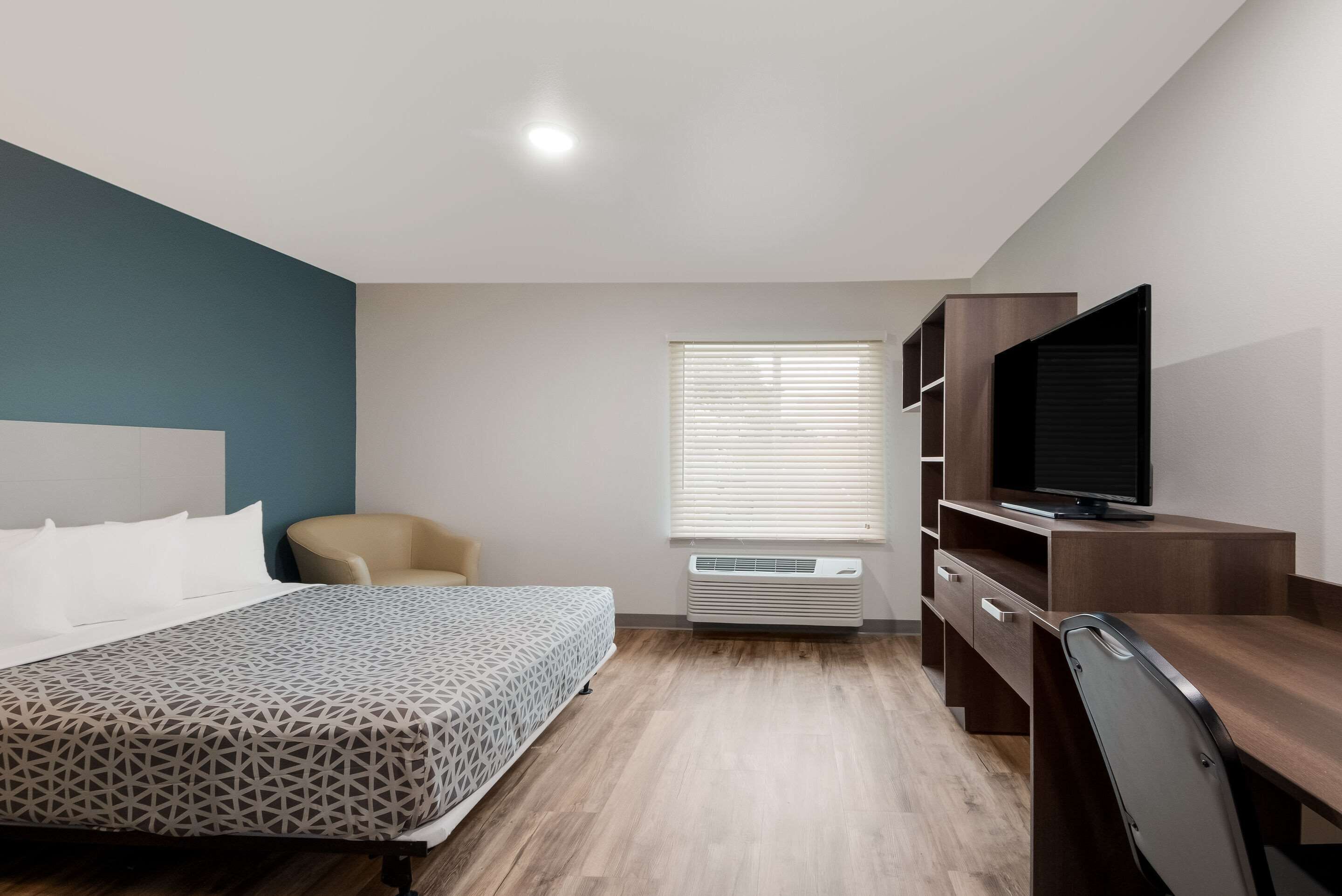 A modern hotel room with a bed, a desk, a chair, a television, and a window with blinds.