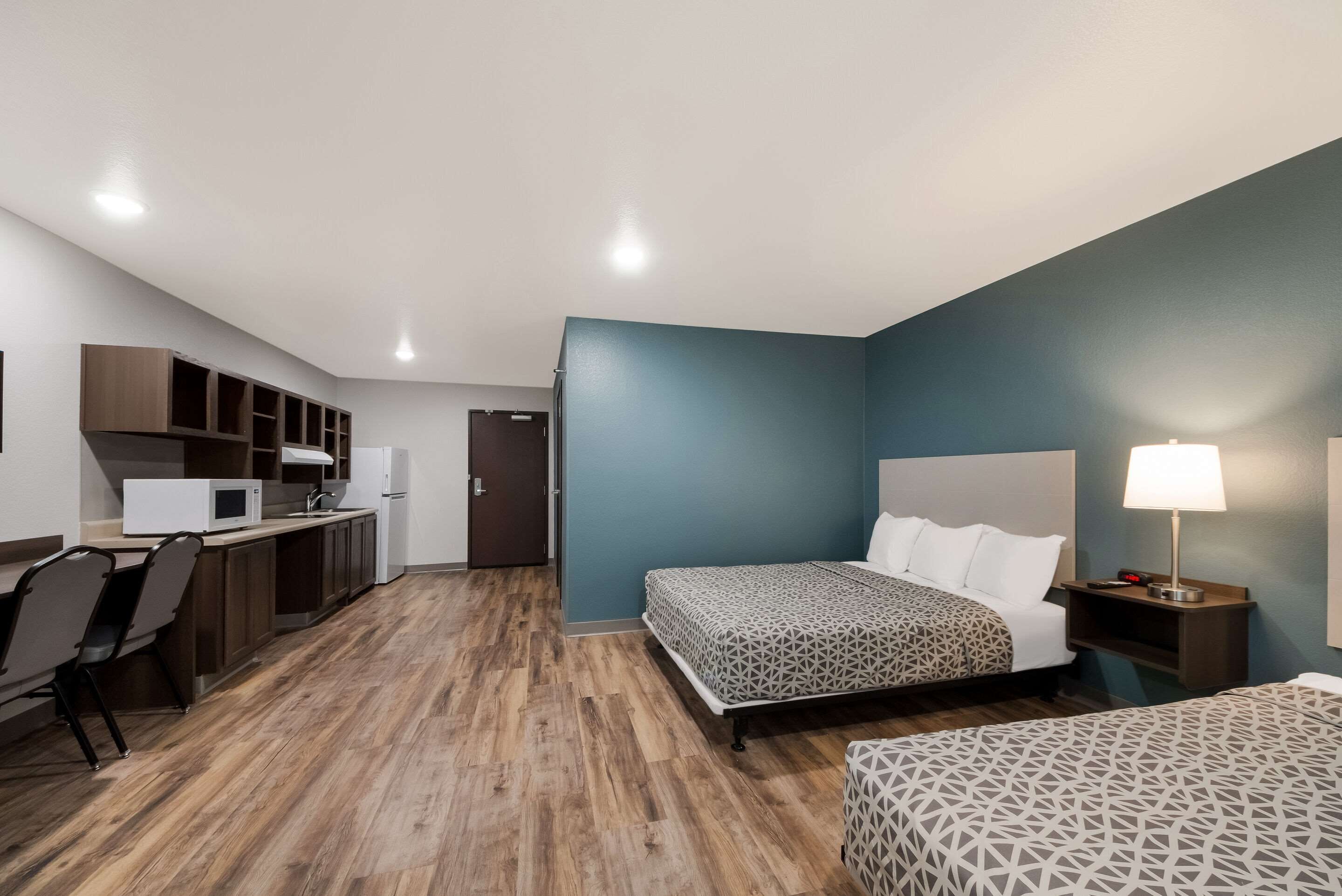 A compact hotel room with a kitchenette, a bed with patterned bedding, and a bedside table with a lamp.