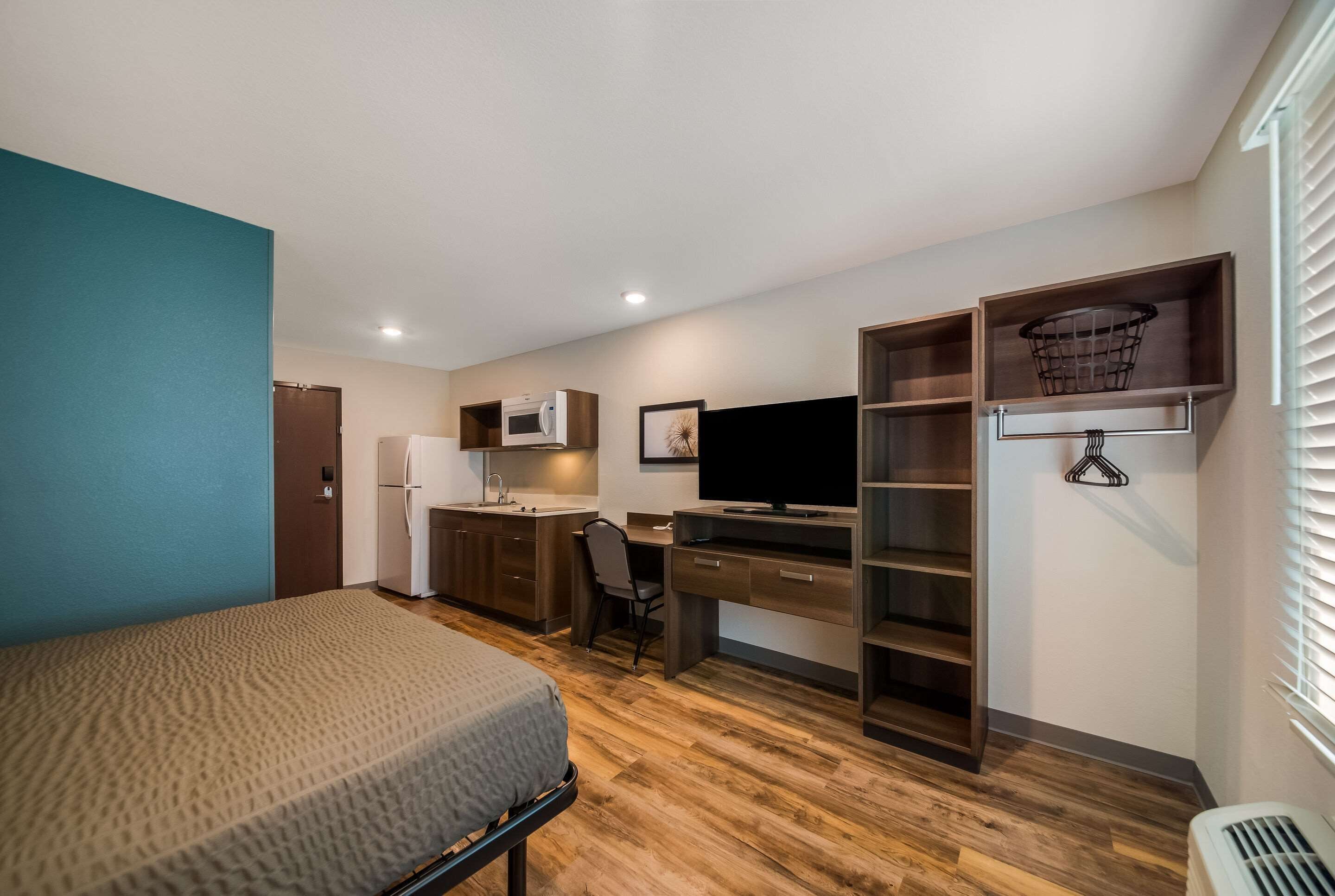 A compact room with a bed, kitchenette, desk, and shelving.