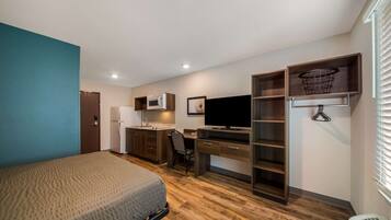 A compact room with a bed, kitchenette, desk, and shelving.