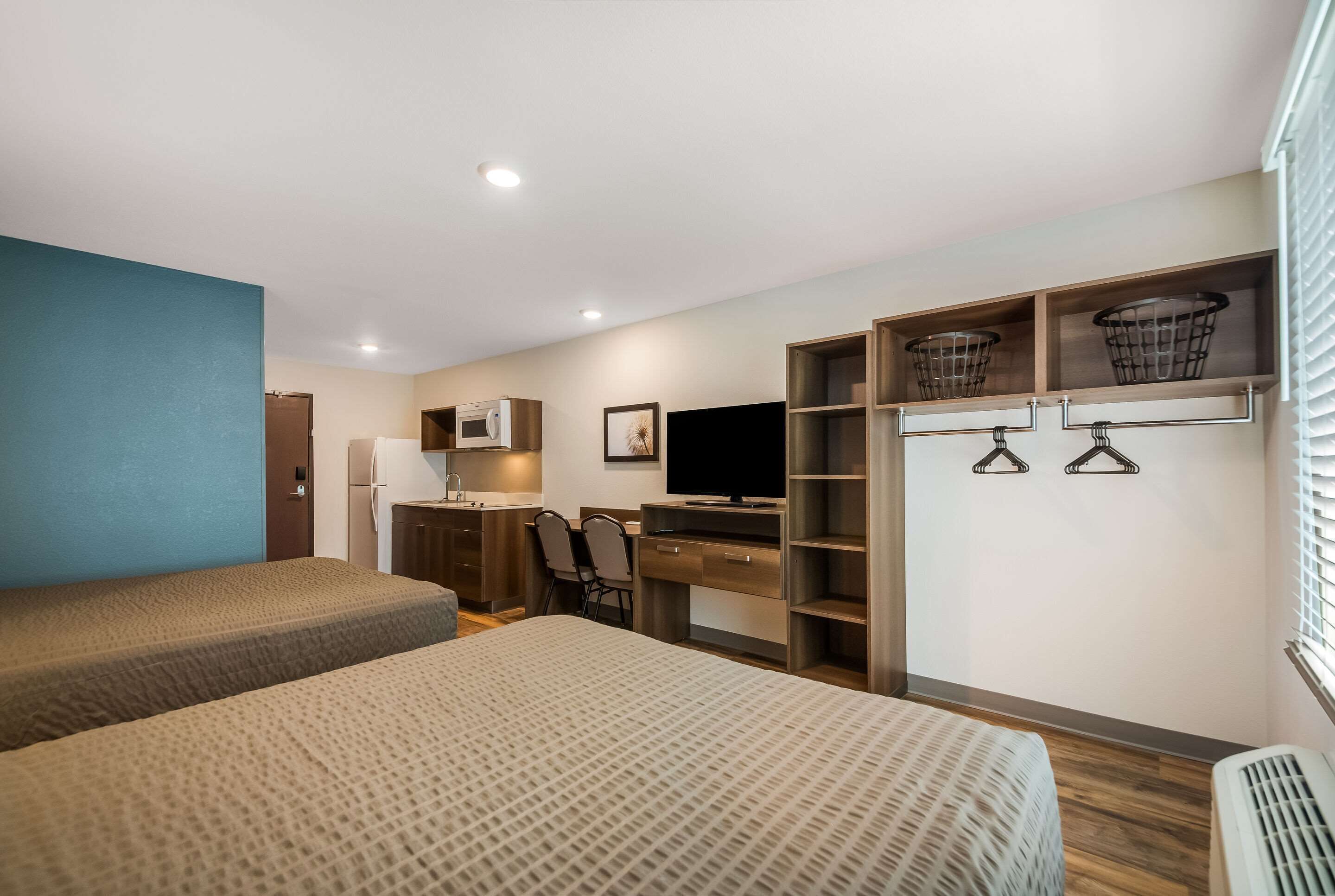 Habitación de hotel con dos camas, un escritorio con silla, televisión y armario con cestas colgadas.