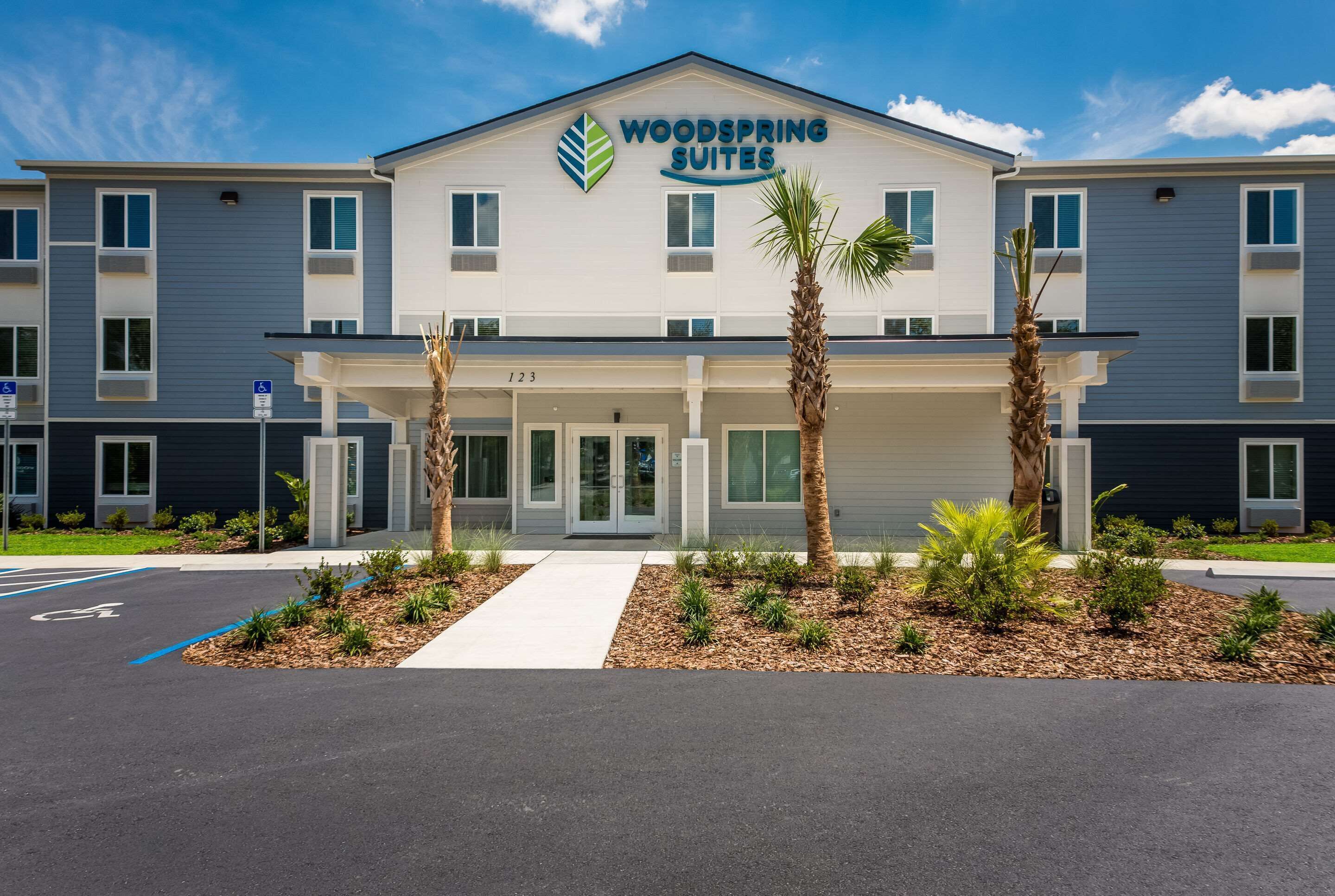 A modern hotel with a sign reading "Woodspring Suites." It features a covered entrance, a parking lot, and a landscaped area with small trees and shrubs.