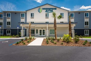 A modern hotel with a sign reading "Woodspring Suites." It features a covered entrance, a parking lot, and a landscaped area with small trees and shrubs.