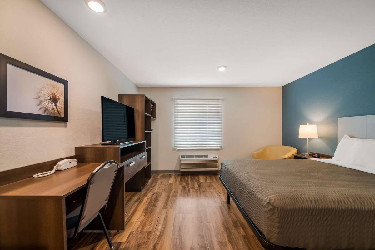 A hotel room with a wooden desk, a bed, a television, a chair, a lamp, and a window with blinds.