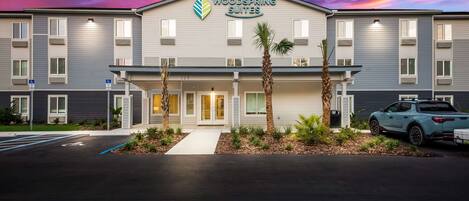 A hotel named Woodspring Suites with a modern design, featuring a flat roof, large windows, and a parking area with a wheelchair-accessible space.