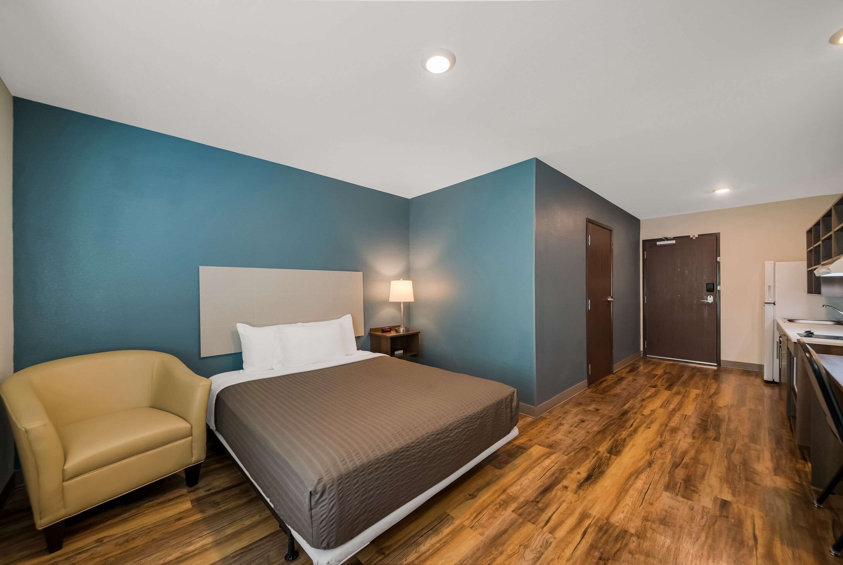 A hotel room with a bed, a chair, a nightstand, a lamp, a wooden floor, a door, and a kitchenette.