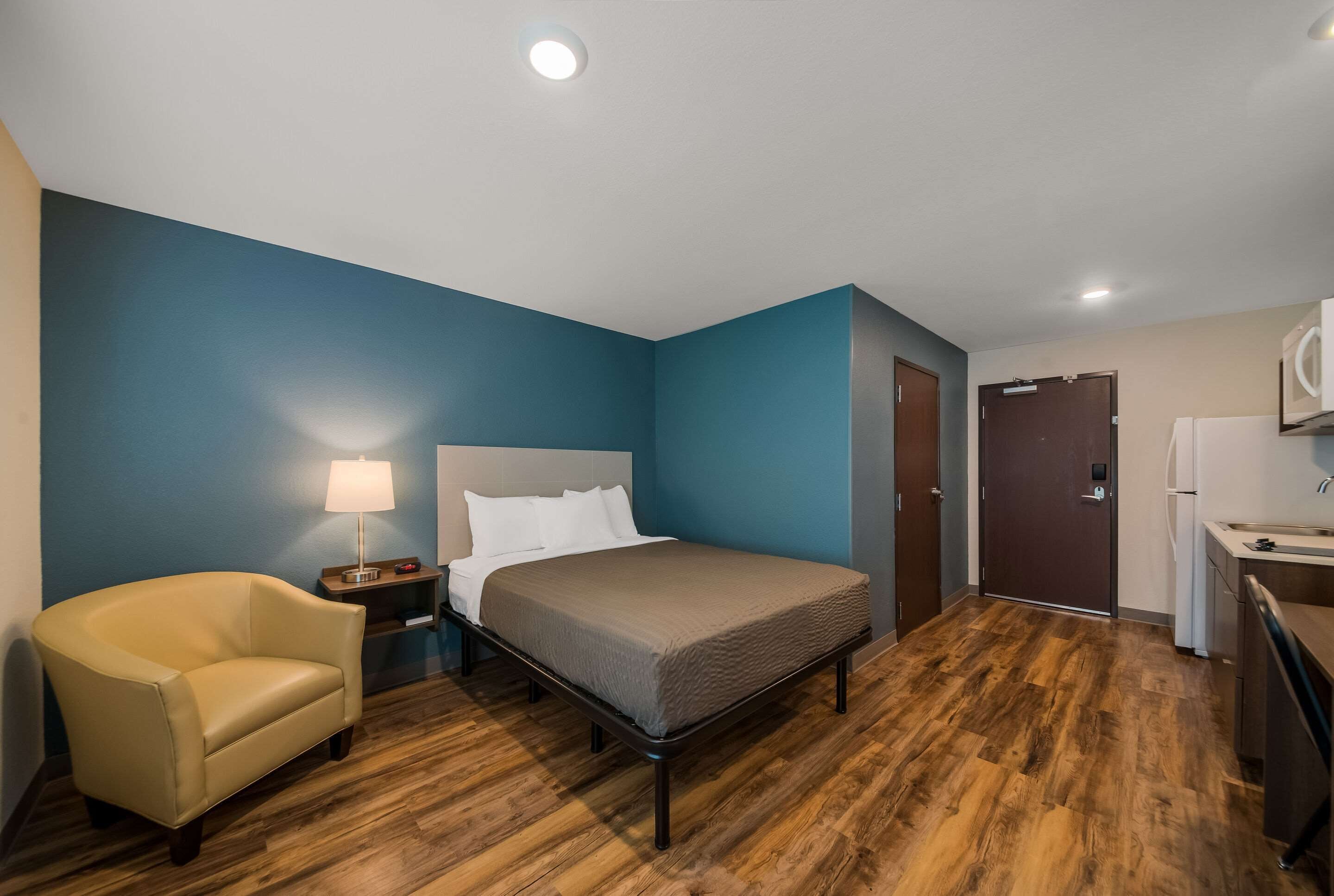 A hotel room with a bed, a chair, a nightstand with a lamp, a wooden floor, a door, and a kitchenette.