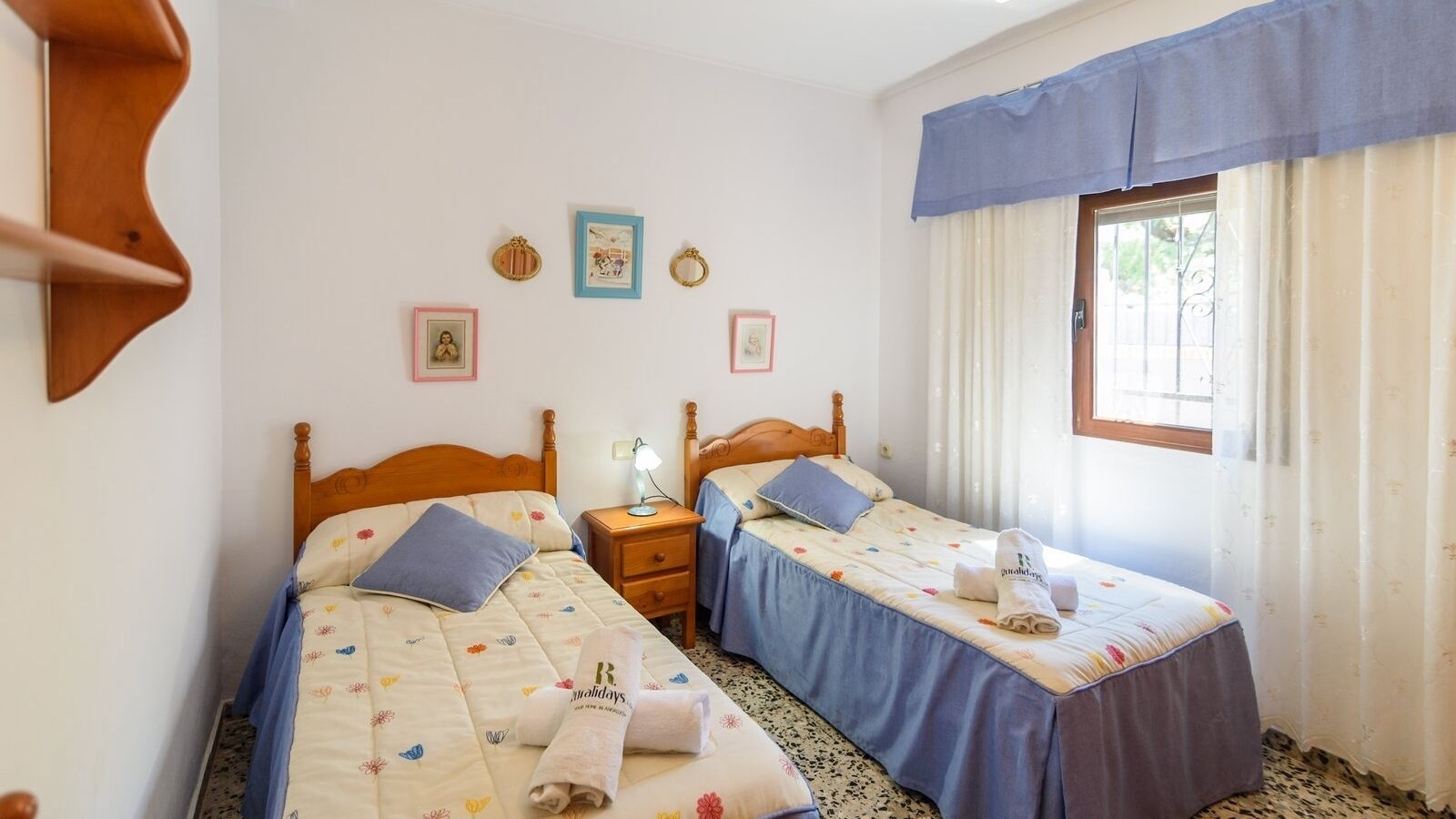 A double room with two single beds, each with a blue bedspread and matching pillows. There are bedside tables with lamps, a wall-mounted shelf, and a window with curtains.