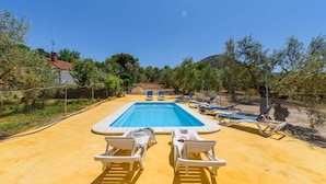 Una piscina rectangular con superficie de agua azul cristalina, rodeada por una zona de arena con sillas de descanso blancas y una valla.