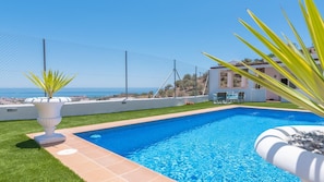 A swimming pool with a clear view of the sea, a white building, and a fenced area with a palm plant.