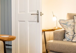 Une pièce avec une porte blanche, une petite table en bois sur laquelle repose une assiette blanche, une lampe sur une table d’appoint en bois, et un canapé gris agrémenté de coussins à motifs.
