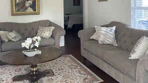 Ein Wohnzimmer mit zwei grauen Sofas, einem Couchtisch mit floralem Teppich, einer Chandelüster und gerahmten Kunstwerken an den Wänden.