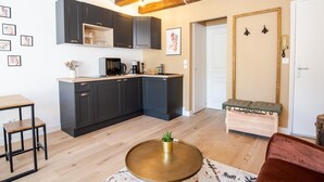 A modern kitchen with dark cabinets, a wooden countertop, and a coffee machine. There is a dining table with chairs and a bench in the living area.