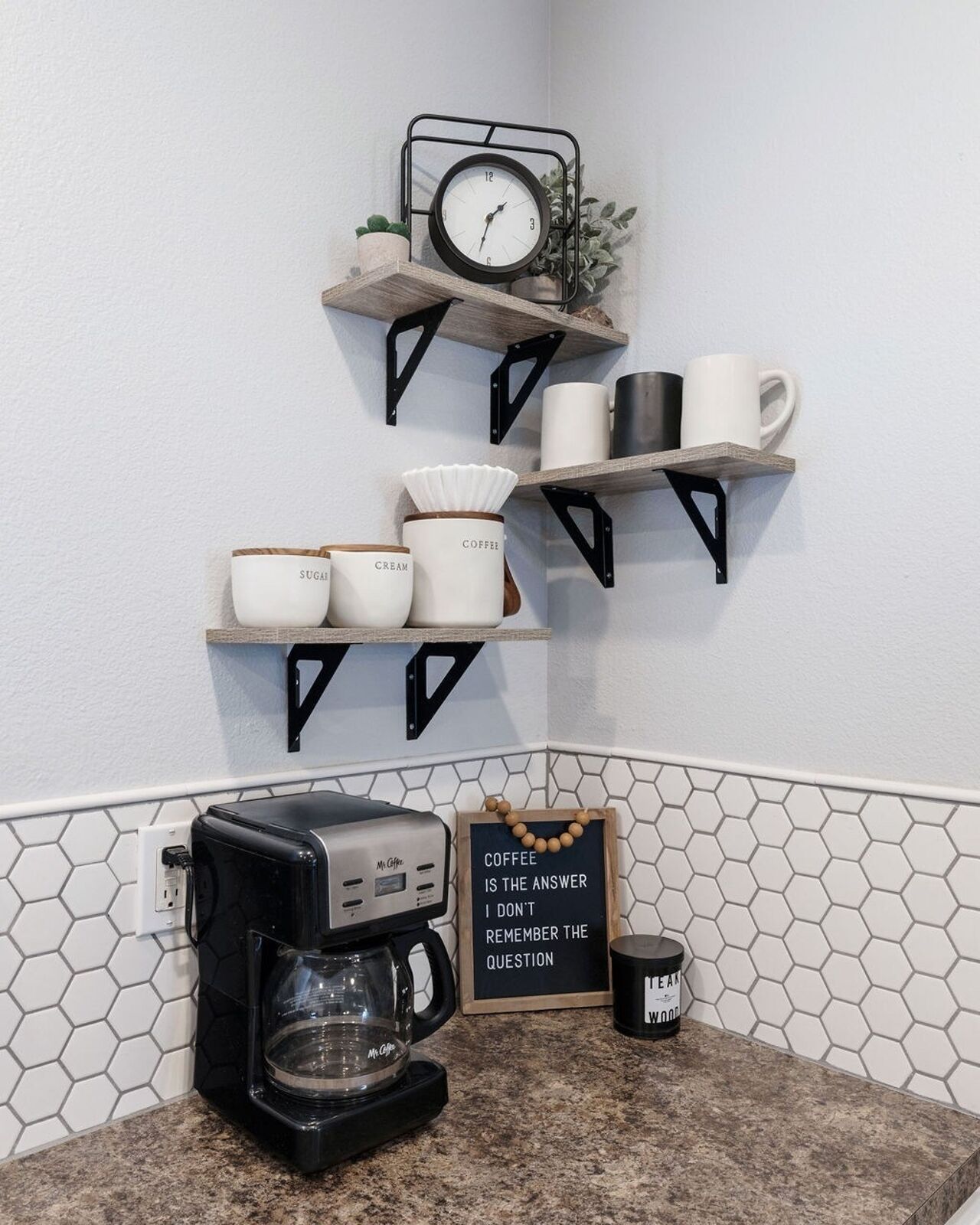 A kitchen with a coffee maker, shelves with mugs and containers, a clock, and a sign with a coffee quote.