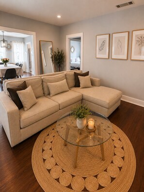 A living room with a sectional sofa, a glass coffee table with a woven placemat, and framed artwork on the wall.